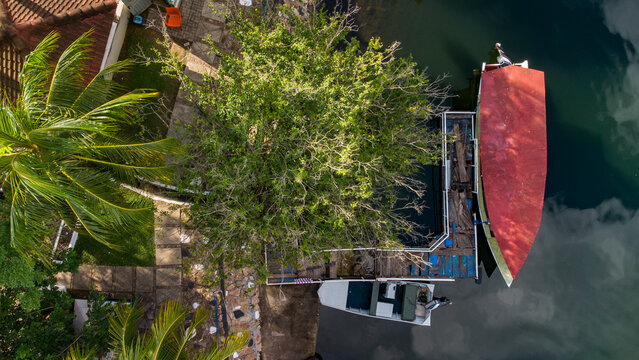 Aerial view of a wooden pier with a boat featuring a large red roof moored on the Volta River, surrounded by lush trees and palm foliage in Atimpoku, Volta Region, Ghana.