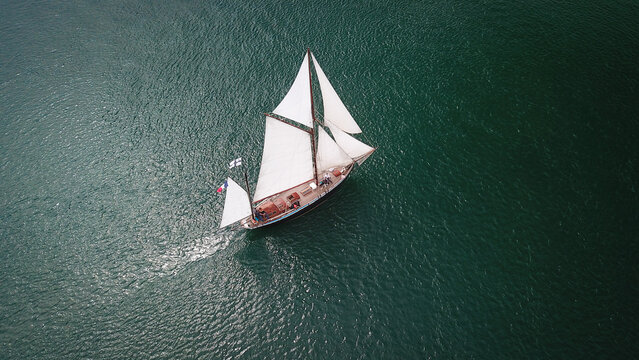 Aerial view of a traditional sailboat with white sails gliding through the calm dark waters of the Golf du Morbihan in Sarzeau, Brittany, France.