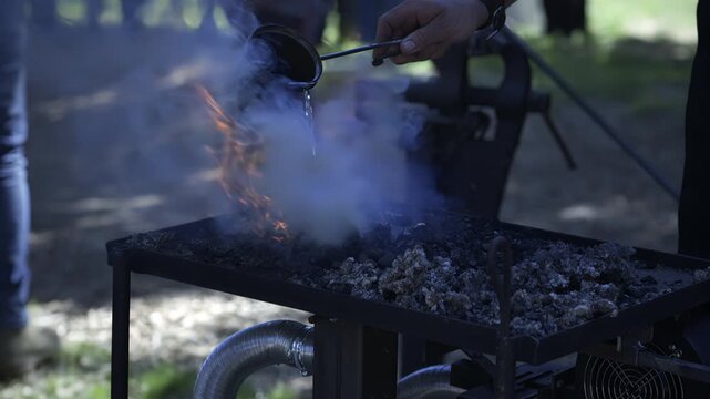 Blacksmith pours water onto a coal forge as thick white steam and smoke rise from the hot fire bed.