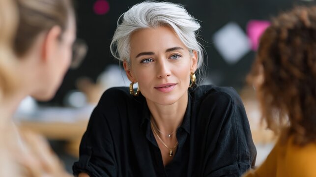 Thoughtful woman with silver hair engaging in conversation at a modern workspace with creative colleagues nearby