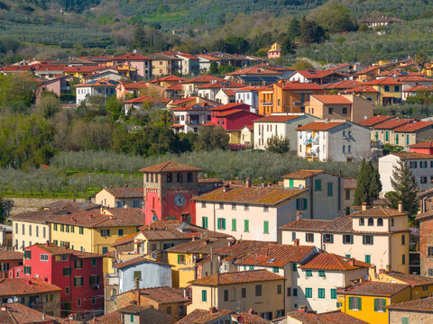 Aerial view of historic colorful buildings with terracotta rooftops, a prominent clock tower, and surrounding olive groves in Loro Ciuffenna, Tuscany, Italy.