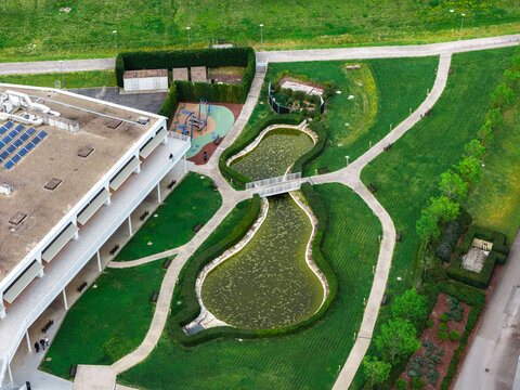 Aerial view of The Mall Luxury Outlets featuring a modern building with solar panels, a landscaped garden with a small pond, walking paths, and manicured hedges in Leccio, Toscana, Italy.