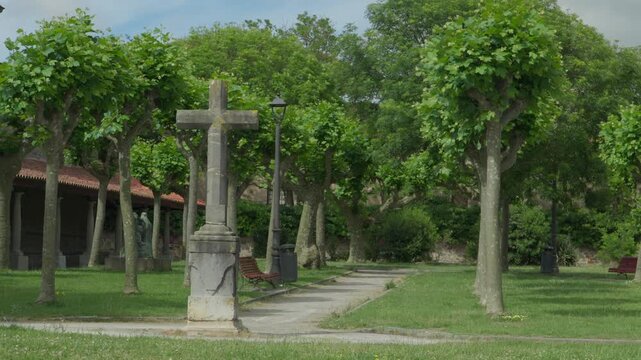 Static shot of a stone cross monument in a tree-lined church garden in Luanco, Spain. Green lawn, lamp post, and historic church arcade visible.