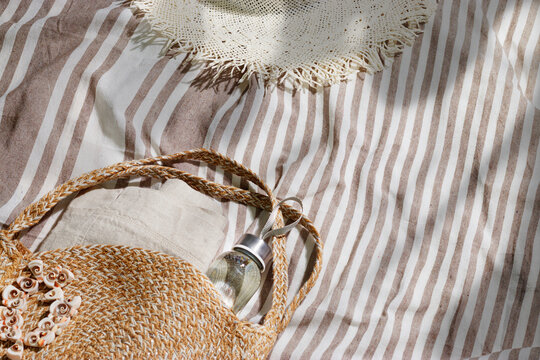Summer background of round crochet bag, bracelet of shell, reusable eco-friendly glass water bottle on striped cloth at sunlight. Sustainable outdoor relaxation and summer lifestyle theme, copyspace.