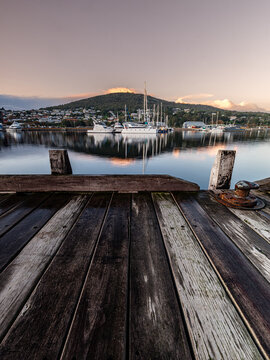 View of Princess Royal Harbour with boats moored in the calm water, reflecting the soft light of dawn, seen from a weathered wooden pier in Albany, WA, Australia.