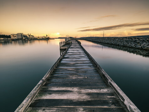 View of the wooden pier extending into the calm waters of Princess Royal Harbour at sunset, with industrial structures visible in the distance in Albany, WA, Australia.