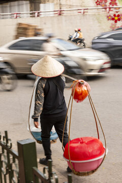 View of a street vendor wearing a traditional conical hat and carrying goods in baskets on a shoulder pole while walking through busy city traffic in Hanoi, Ha Noi, Vietnam.