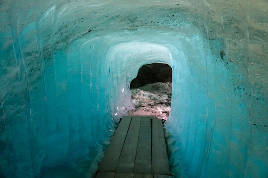 View of the ice tunnel inside the Rhone Glacier with a wooden walkway and vibrant blue ice walls in Obergoms, Valais, Switzerland.