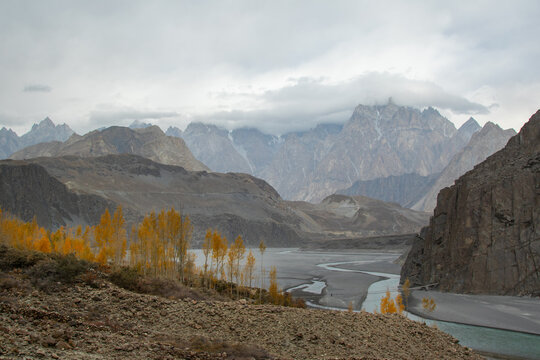 View of the Passu Cones mountain range with golden autumn trees along the riverbank under a cloudy sky in Hunza Nagar, Gilgit, Pakistan.