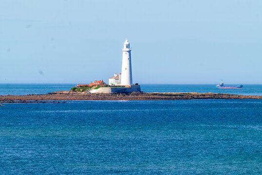 View of St Mary's Lighthouse on a rocky island surrounded by the North Sea under a clear blue sky with a cargo ship in the distance in Whitley Bay, England, United Kingdom.