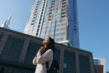 Fototapeta premium Woman gazing upward in an urban canyon of glass and steel, hands in prayer near a modern skyline, capturing hope, focus, and contemplation amid tall buildings and clear blue sky.