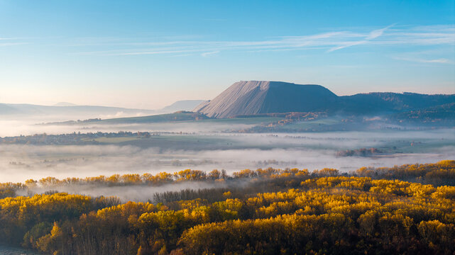 Aerial view of Monte Kali salt mountain rising above a misty valley with autumnal yellow trees under a clear blue sky in Heringen, Hessen, Germany.