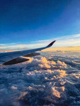 Aerial view of an airplane wing with a red wingtip flying over a thick layer of cumulus clouds illuminated by the golden light of sunset under a clear blue sky in Africa.