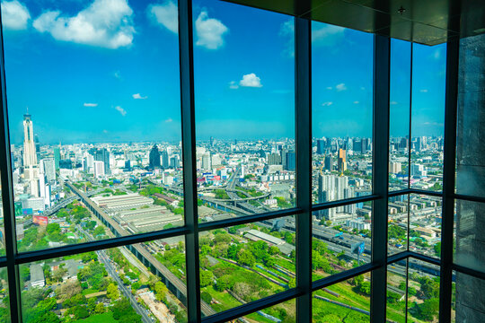 Aerial view of Bangkok, Thailand, seen through a glass window with dark framing, showing the skyline, rail lines, highways, and green urban spaces under a bright blue sky.