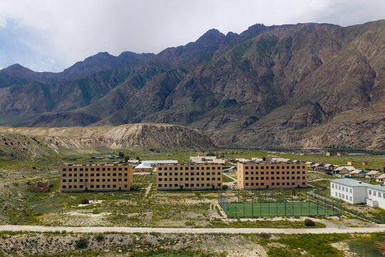 Aerial view of abandoned Soviet-era apartment buildings and a green sports field at the foot of rugged mountains in Enilchek, Issyk-Kul Region, Kyrgyzstan.