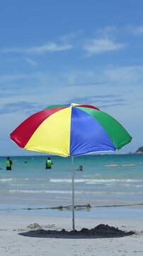 Beach umbrella on beach with sea and sand, summer vacation travel under blue sky, colorful parasol casting shadow on wet shoreline, gentle waves, distant swimmers, copy space