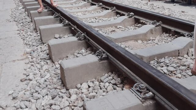 Feet wearing sandals standing on railway track sleepers and gravel ballast, representing travel, waiting, or exploring transportation infrastructure in a tourist destination with other people nearby