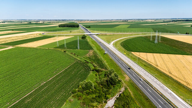 Aerial view of a highway stretching through vast agricultural fields and green farmlands under a clear sky in Sremska Mitrovica, Serbia.