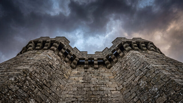 Majestic weathered stone castle tower featuring thick defensive walls and ornate battlements captured from a low angle perspective against a dramatic moody sky filled with dark rolling gray storm clou