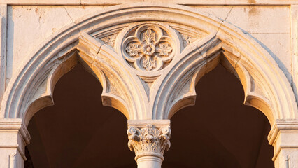 Obraz premium Intricate stone carving featuring a delicate quatrefoil floral motif centered above a decorative column capital on a historic limestone archway illuminated by the warm golden glow of the afternoon sun