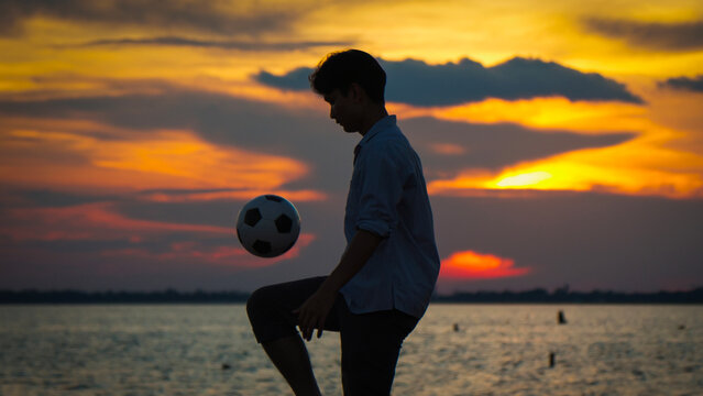 Silhouette of a young man juggling a soccer ball by the lakeside at sunset. Lifestyle
