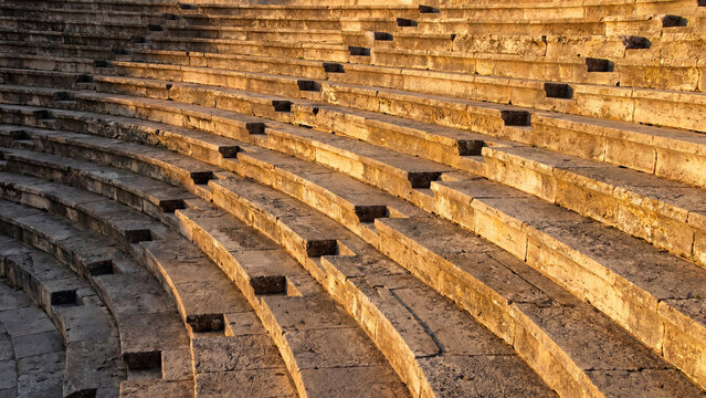 Ancient stone tiered seating rows of a historic mediterranean amphitheater glowing under the warm golden light of a late afternoon sun during a peaceful day at an archaeological ruin site.