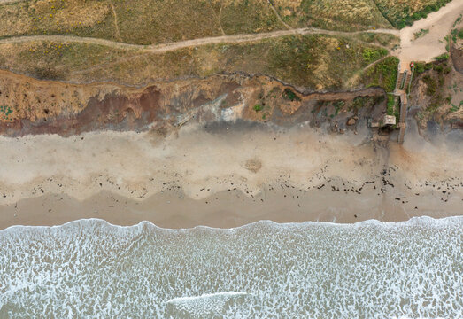 Aerial view of the sandy shoreline and eroded cliffs of Compton Bay with waves crashing on the beach and a wooden staircase in Newport, Isle of Wight, United Kingdom.