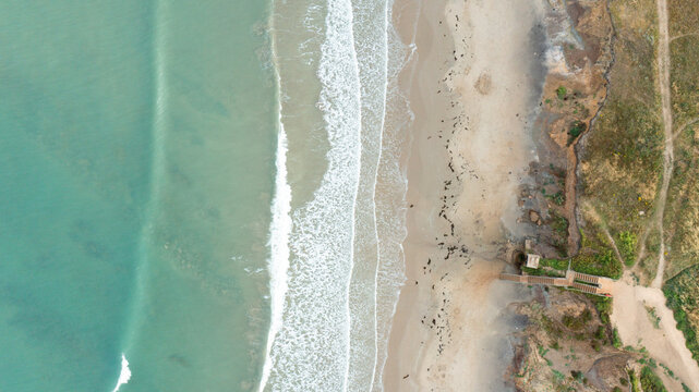 Aerial view of the turquoise waves of Compton Bay crashing against the sandy shore and wooden staircase at Newport, Isle of Wight, United Kingdom.