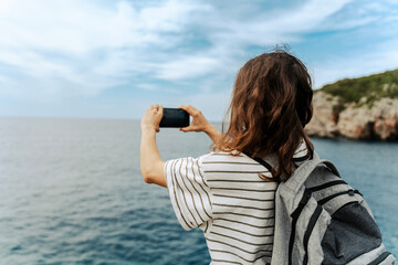 Rear view of a female tourist with a backpack taking a photo of a scenic sea landscape with her...
