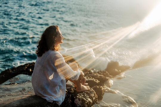 Young woman sitting on a stone rock facing the sea in a bright sun flare and enjoying summer