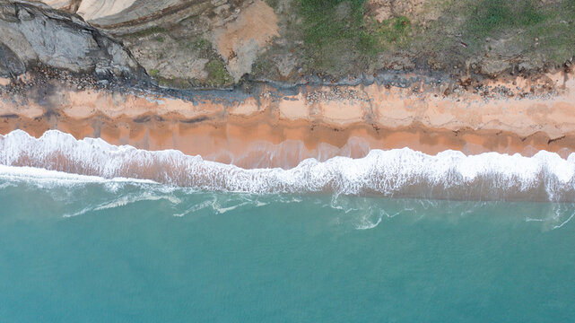 Aerial view of the sandy shoreline and turquoise waves crashing against the base of the steep cliffs at Whale Chine, Ventnor, Isle of Wight, United Kingdom.