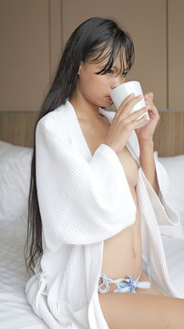 Attractive young Asian woman in an open white bathrobe sitting on a bed and drinking coffee from a white mug