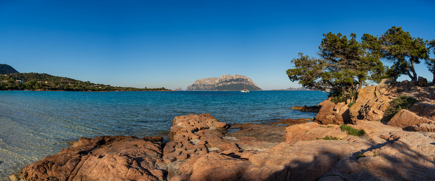 View of Porto Istana Beach with its characteristic red granite rocks, turquoise sea, and the distant Tavolara island under a clear blue sky in Sardinia, Italy.