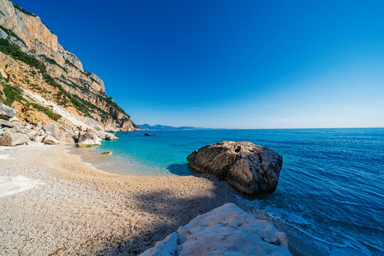 View of the turquoise waters and white pebble beach of Cala Goloritze, framed by dramatic limestone cliffs under a clear blue sky in Sardinia, Italy.