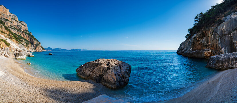 View of the turquoise waters and white pebble beach of Cala Goloritze, framed by dramatic limestone cliffs under a clear blue sky, Sardinia, Italy.