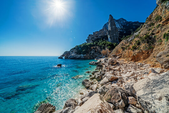 View of Cala Goloritze with its iconic limestone pinnacle, turquoise waters, and rocky shoreline under a bright sun in Sardinia, Italy.