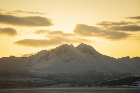 View of the snow-covered mountains of Tromso under a golden sunset sky in Tromso, Troms, Norway.