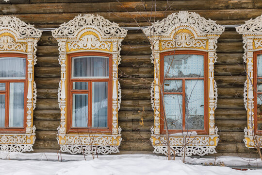View of traditional wooden house with intricate white carved window frames on a log wall in winter with snow on the ground in Tomsk, Tomsk Oblast, Russia.