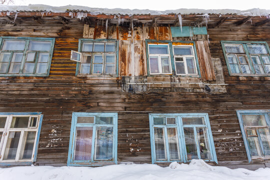 View of the facade of a weathered wooden house with blue window frames, peeling paint, and icicles hanging from the roof in winter in Tomsk, Tomsk Oblast, Russia.