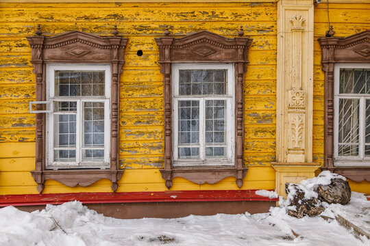 View of the weathered yellow wooden facade of a historic house with ornate carved window frames and snow on the ground in Tomsk, Tomsk Oblast, Russia.