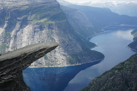 View of the Trolltunga rock formation overlooking the deep blue waters of Ringedalsvatnet lake, surrounded by steep mountain cliffs and glaciers in Ullensvang, Vestland, Norway.
