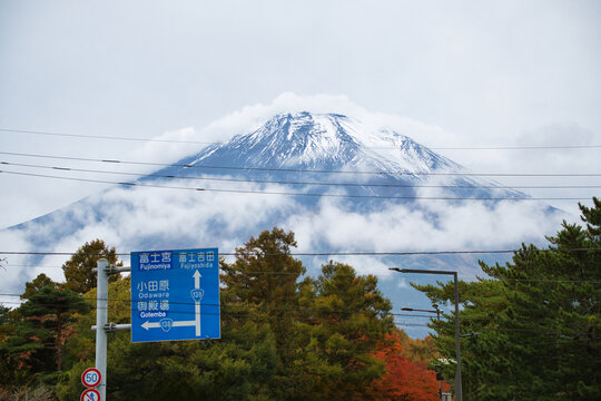 View of Mount Fuji with snow-capped peak partially obscured by clouds, framed by autumn foliage and a blue road sign in Fujinomiya, Shizuoka, Japan.