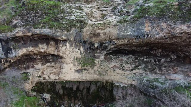 Aerial shot flying close to a monumental limestone cliff featuring layered cavern alcoves, a dark grotto entrance and fresh moss patches in Kazakhstan. 