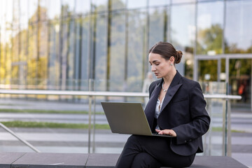 Fototapeta premium Businesswoman works on laptop outside modern office building during daylight hours in urban area