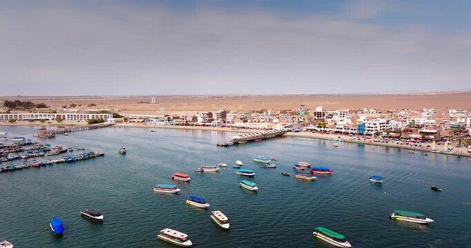 Numerous colorful boats anchored in calm ocean water near pier and desert town in Paracas, Peru. Aerial backward, copy space