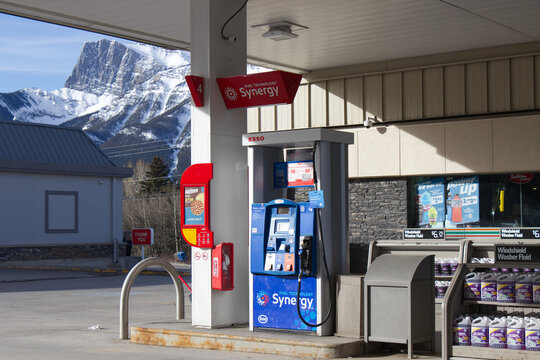 An Esso Synergy fuel pump and convenience store with a windshield washer fluid display stand below a snow-capped mountain at a gas station on a clear spring morning.