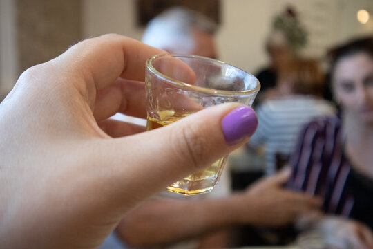Mano di donna con unghie laccate di viola che tiene un bicchierino da shot pieno di liquore ambrato davanti a un muro di pietra rustica