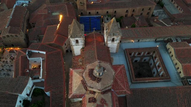 Top down drone flyover above a historic church complex with cloister courtyard and illuminated towers at dusk in old town Spain.