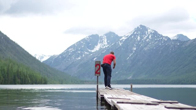 A male adventurer in a red t-shirt skipping stones across the calm water surface of Multinskoe Lake against a snow-capped mountain backdrop.