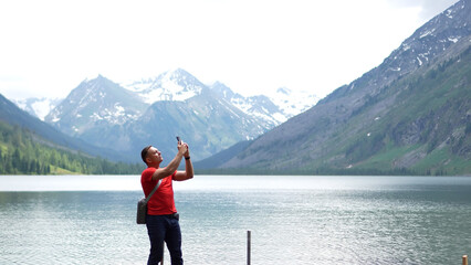 Fototapeta premium An athletic male adventurer in a red t-shirt standing on a lake shore and using his smartphone to take a photo of distant snowy mountain peaks.